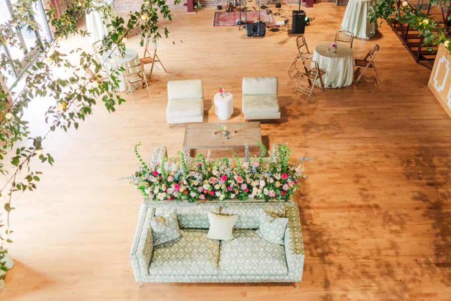 Overhead view of Power House venue with hardwood floors, greenery on market lights, lush, bright flowers, beautiful sage green lounge furniture and bamboo chairs.