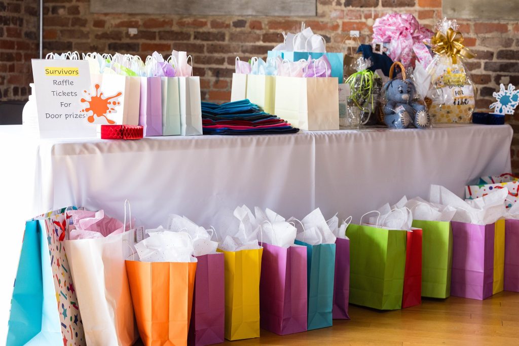 Colorful gift bags and baskets cover a table and are also lined up on the floor at an event at the Power House.