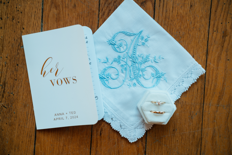 The bride's vow book lies next to a white handkerchief embroidered with the bride's initial in blue. Her wedding band and engagement ring sit in a white ring box next to it.