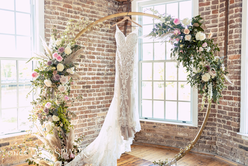 A white sleeveless lace wedding dress hangs on a circular arbor, adorned with beautiful pink and white flowers, with lush greenery. The arbor is set in the River Overlook of the Power House at Rocky Mount Mills.