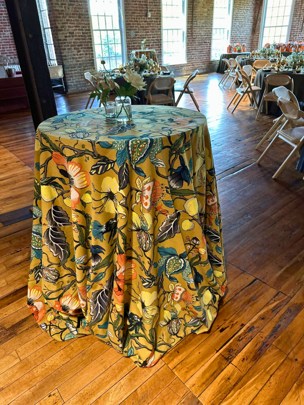 A cocktail table covered with vibrant, floral linen in shades of ochre, teal, yellow and orange, in the River Overlook of the Power House. Guest tables are in the background.