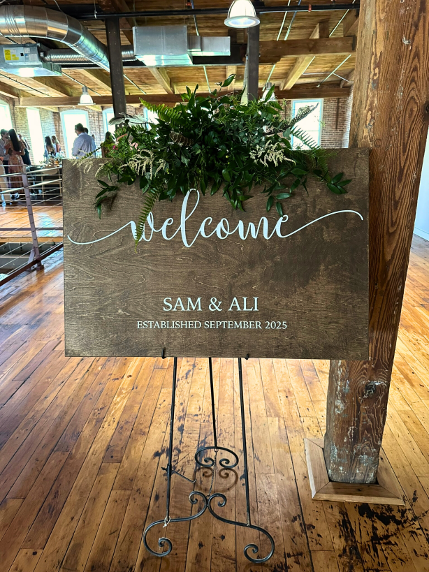 A wooden sign that says "Welcome: Sam & Ali, Established September 2025" topped with greenery.