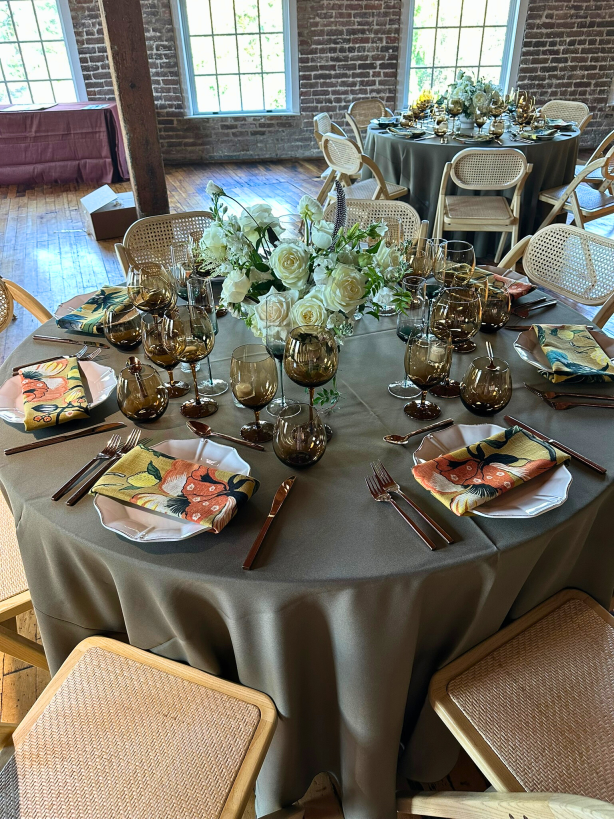 A table setting with amber glassware, white florals, bright patterned napkins, gold flatware on a table with a moss colored linen.