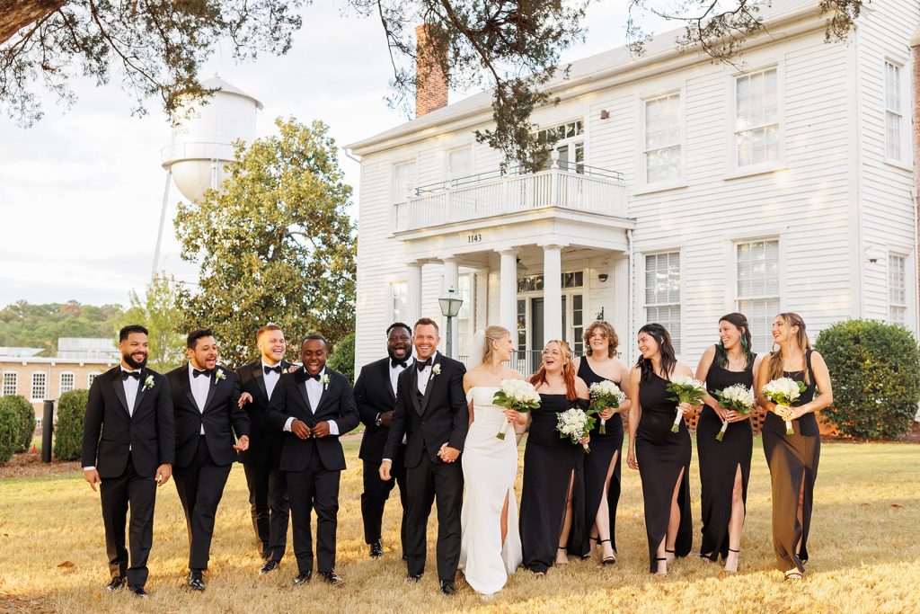 The bride and groom walk towards the camera with their bridal party, in front of a white house.