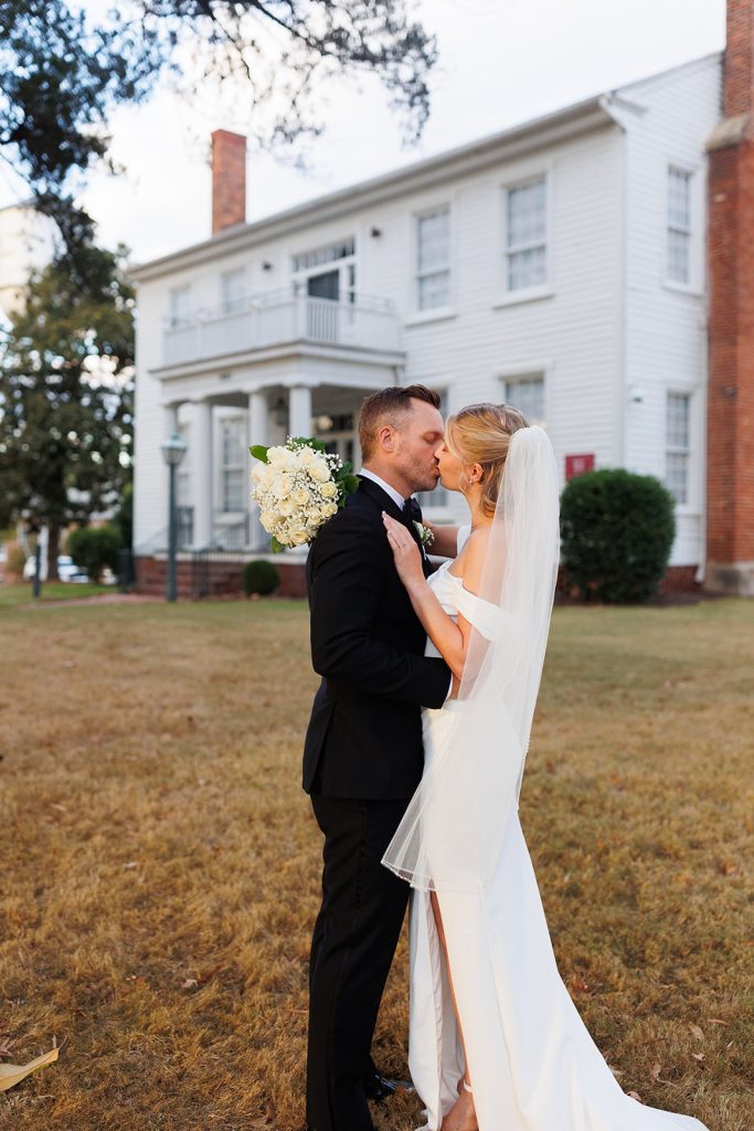 Bride and groom share a kiss in front of a white house.