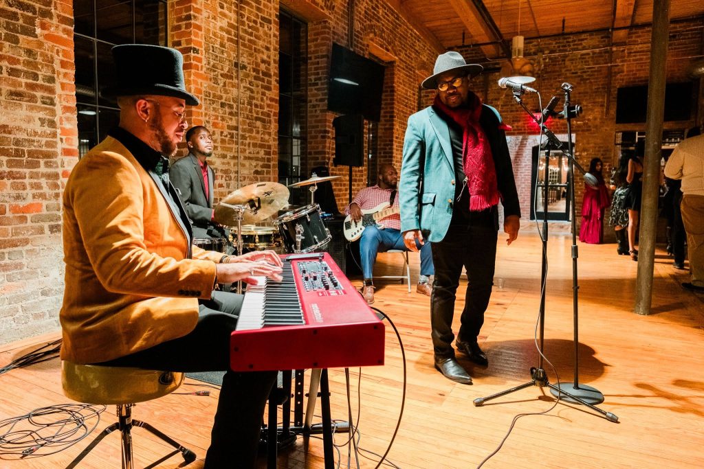 A 3 piece band plays on the Switchgear level of the Power House at Rocky Mount Mills. The singer is pausing for a keyboard solo during the performance at a corporate Christmas party.