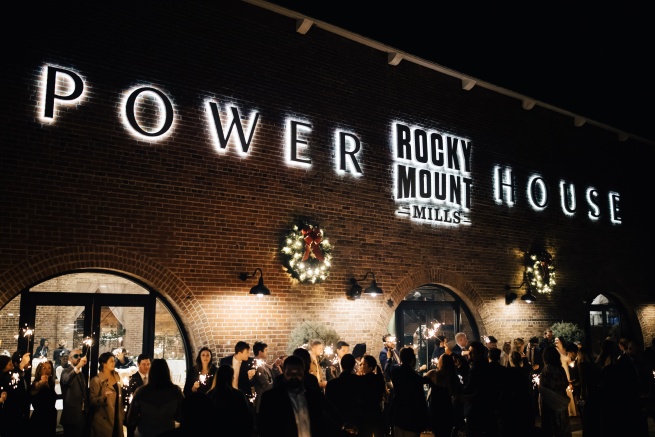 The exterior of the Power House at Rocky Mount Mills is illuminated, with lighted Christmas wreaths. A crowd of people gather outside with sparklers. Photo Credit: Amanda Sutton Photography